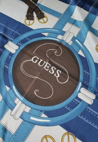 Tessuto a righe blu e bianche con emblema circolare marrone incorniciato da anelli blu, che presenta la parola "GUESS" e cinture in pelle illustrate con fibbie dorate.