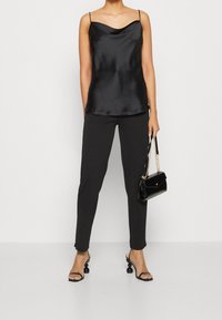 Camisole en satin noir avec décolleté drapé, pantalon noir ajusté, et sandales à lanières avec talons ronds. Petit sac à main noir avec chaîne.