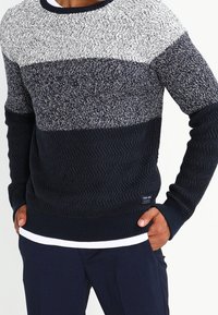 Homme portant un pull tricoté à blocs de couleurs bleu marine, gris et blanc avec des motifs texturés et un pantalon bleu marine, mains dans les poches.