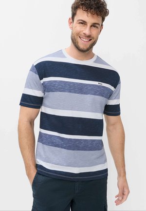 Lächelnder Mann trägt ein kurzärmeliges T-Shirt mit horizontalen marineblauen, blauen und weißen Streifen und steht mit einer Hand in der Tasche vor einem schlichten Hintergrund.