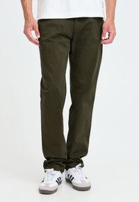 Homme portant un pantalon décontracté vert foncé et des baskets blanches avec des rayures noires, debout devant un fond blanc uni.