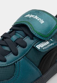 Zapatilla deportiva de cuero color verde azulado con detalles en negro, que presenta una superposición texturizada, diseño con cordones y una correa de velcro con detalle de logo.