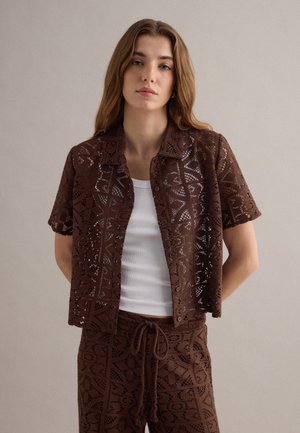 Giovane donna che indossa una camicia marrone a maniche corte con fantasia e pantaloni abbinati sopra una canottiera bianca senza maniche, in piedi davanti a uno sfondo semplice.