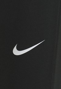 Leggings de sport noirs en tissu extensible, avec un logo Nike en argent sur le côté et des détails de couture visibles.