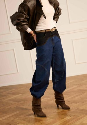 Persoon met een bruine leren jas, wit shirt, high waist spijkerbroek in twee tinten blauw en bruine laarzen met hak, staande op een houten vloer.