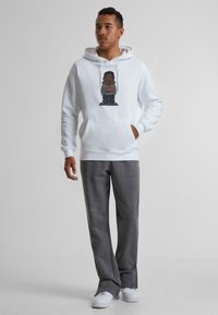 Hvid hættetrøje med grafisk design i midten, grå bukser og hvide sneakers. Hættetrøjen har en forlommekasse og snore i hætten.