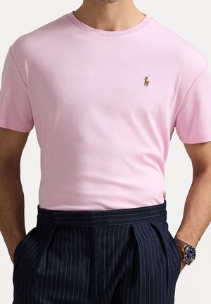 Mężczyzna ubrany w jasno różowy krótki rękaw T-shirt z małym haftowanym logo polo oraz ciemne spodnie w cienkie prążki.