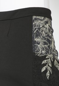 Jupe noire avec panneau latéral orné de broderies florales blanches et motifs texturés, taille ajustée et détail plissé subtil à l'avant.