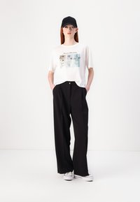 White t-shirt with floral print and text "The art of Blooming," paired with loose black trousers and white sneakers. Black cap on head.