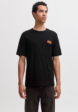 Homme portant un t-shirt noir Jack & Jones avec un patch logo orange, un pantalon foncé et un collier chaîne argenté, debout devant un fond clair uni.