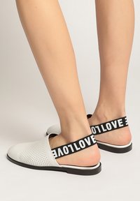 Baskets blanches à enfiler avec un design perforé, sangle élastique noire comportant le texte "LOVE". Bout arrondi et semelle plate avec un minimum de détails.