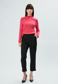 Enganliegender pullover in Hotpink mit Zopfmuster, kombiniert mit schwarzen, tapered Hosen und schwarzen, spitzen Pumps.