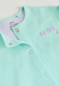 Body bébé côtelé vert menthe avec boutons-pression, texte « Smiles » en violet et visage souriant rose sur la poitrine, col intérieur violet clair.