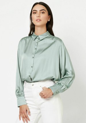 Satingrüne Bluse mit klassischem Kragen und Knopfverschluss, ausgestattet mit langen Ärmeln und gesammelten Manschetten, getragen zu weißen, hoch taillierten Hosen.