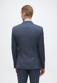 Casaco de fato azul escuro feito de tecido texturizado, com uma fenda única, lapelas em bico e punhos com botões. Usado sobre calças a condizer.