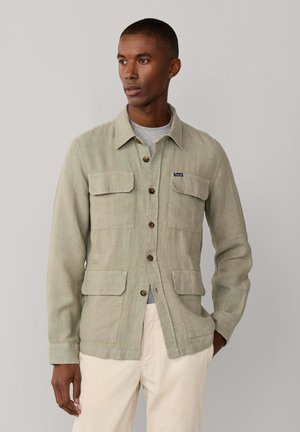 Hombre con chaqueta de lino verde claro abotonada con cuatro bolsillos con solapa, camiseta gris y pantalones color crema sobre un fondo liso.