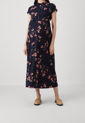 Femme enceinte portant une robe de maternité florale bleu marine à manches courtes, col, et des chaussures plates camel, debout devant un fond uni.