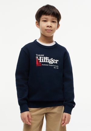 Dreng iført navyblå Tommy Hilfiger sweatshirt med rød sejlbåd-logo og beige bukser, står foran en ensfarvet hvid baggrund.