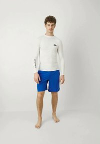 Baltā garpiedurkņu aizsargkrekls ar melnu logotipu uz krūtīm un piedurknēm, savienots ar vienkrāsainiem zils peldbiksēm. Modelis ir basām kājām.