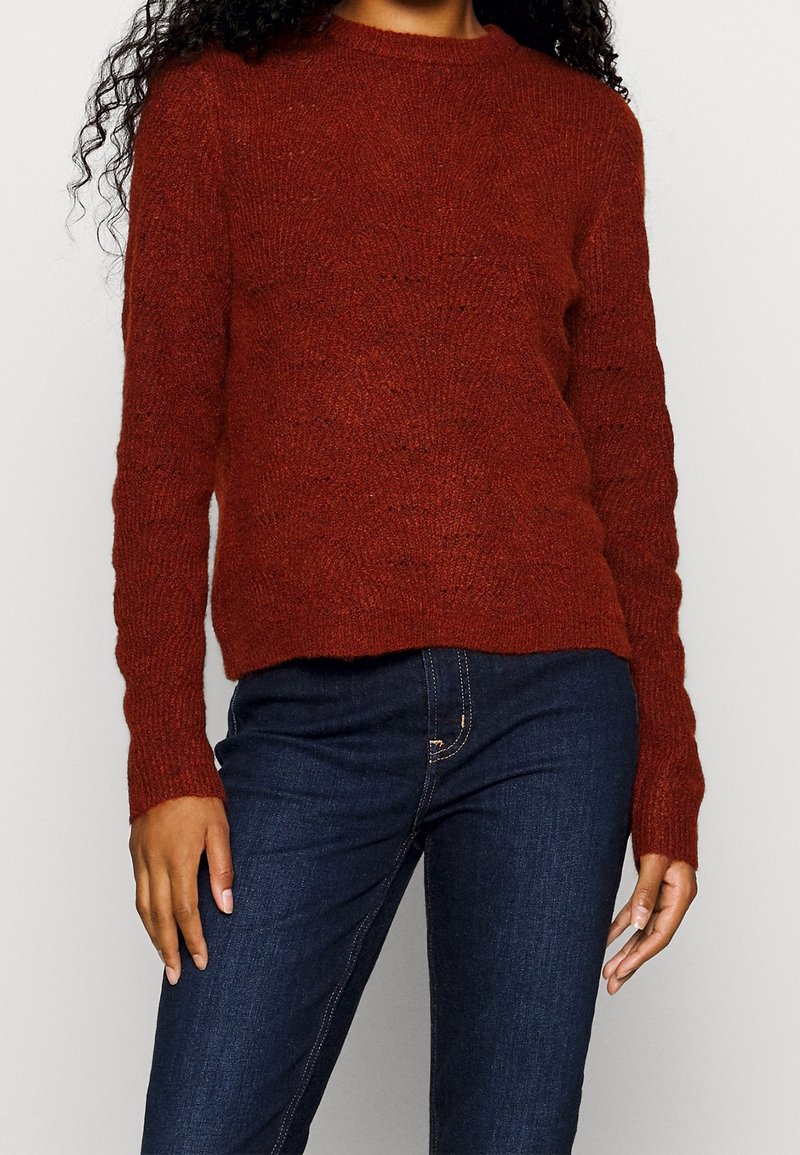 Femme portant un pull en maille couleur rouille à manches longues et un jean slim bleu foncé sur un fond clair uni.