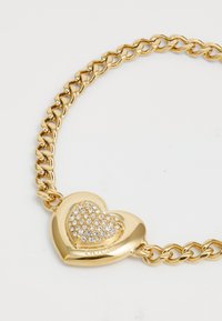 Bracelet en chaîne dorée avec un charm en forme de cœur orné de strass transparents. Le charm possède un nom de marque gravé et une finition lisse et brillante.