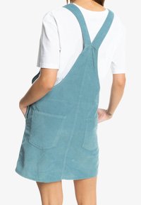 Modré kordové šaty na celkovou sukni s dvěma předními kapsami, nastavitelnými ramínky a jemnou strukturou. Mají délku těsně nad kolena.