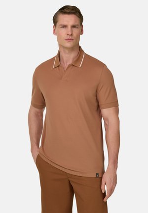 Homme portant un polo marron à manches courtes avec un liseré blanc sur le col et un pantalon marron assorti, debout avec une main dans la poche.