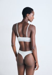 Kvinne med kort krøllete hår som har på seg en lys grå Tommy Hilfiger-bh og matchende truse, sett bakfra mot en ensfarget hvit bakgrunn.