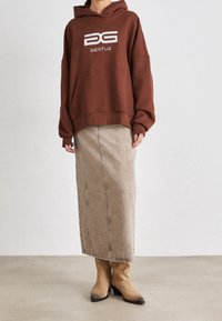 Osoba oblečená v hnědém oversized mikině s logem "GESTUZ", béžové dlouhé sukni a světlých sukněních botách stojí na obyčejné podlaze.