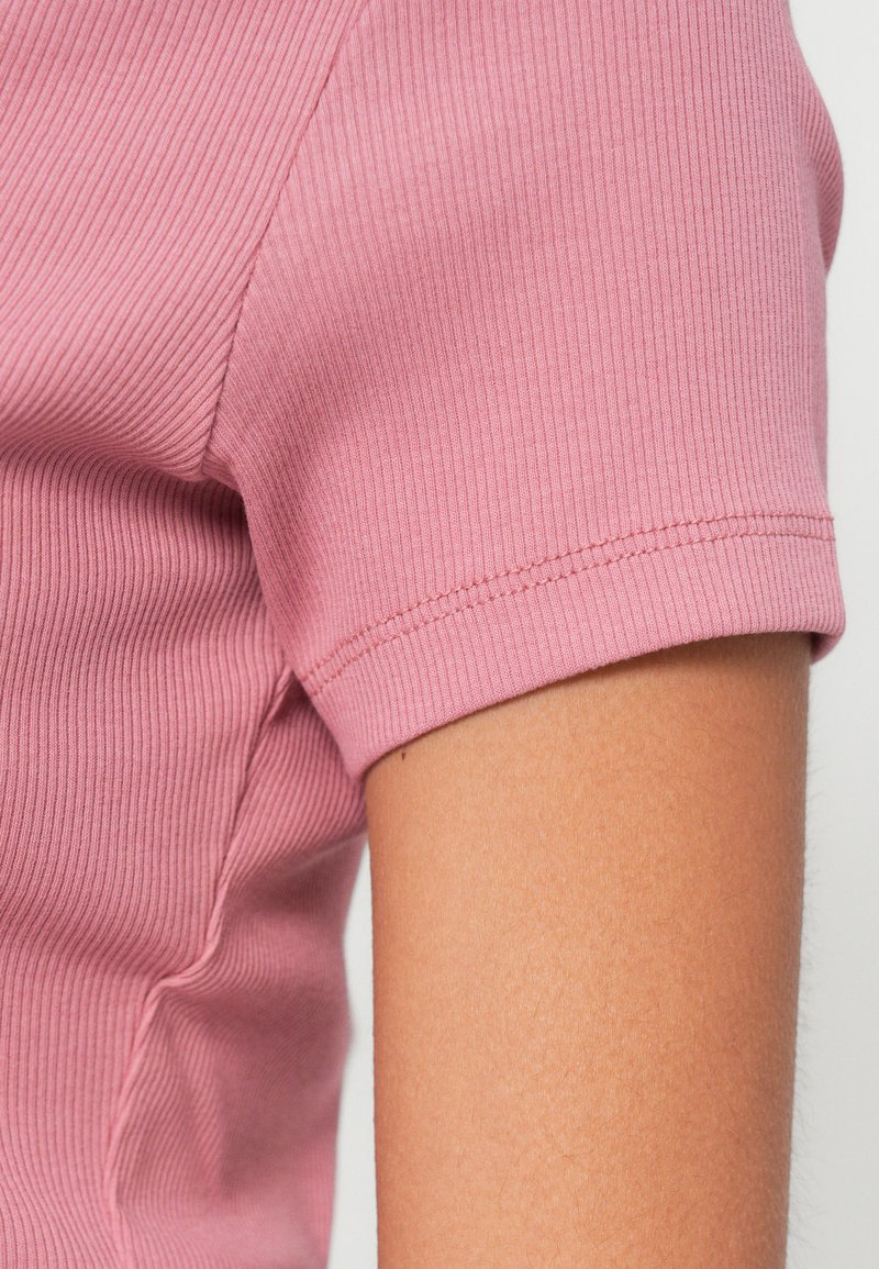 Hellrosa geripptes Kurzarmshirt mit enger Passform; glatte Textur mit sichtbaren Nahtdetails entlang des umgeschlagenen Ärmelsaums.