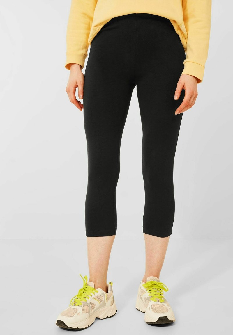 Black cropped leggings with a smooth texture and elastic waistband, paired with light-colored sneakers featuring bright green laces.