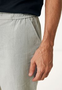 Ljust grå linnebyxor med elastisk midja, med en sidoficka och ett slätt, strukturerat tyg. Hand synlig på sidan.