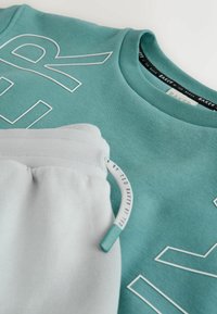 Miętowa bluza z białym napisem, w połączeniu z jasnoszarymi spodniami dresowymi. Oba wykonane z miękkiego materiału, z elastycznym paskiem w talii i detalem z metką.