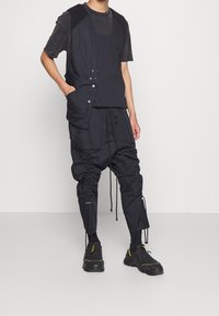 Schwarzes, geschichtetes Outfit mit einem Weste mit Knöpfen und Taschen, kombiniert mit lockeren, gerafften Hosen. Zusätzlich schwarze Schuhe mit gelben Akzenten.