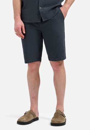 Man die een textuurrijke donkere korte broek en bijpassend overhemd met korte mouwen draagt, met bruine suède instappers, staand met ontspannen handen.