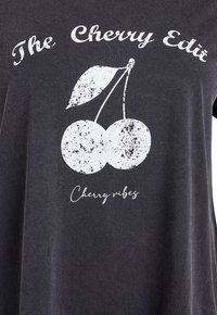 Schwarzes Hemd mit weißem, verwaschenem Kirsch-Grafikdruck und Text "The Cherry Edit" oberhalb sowie "Cherry vibes" unterhalb der Kirschen.