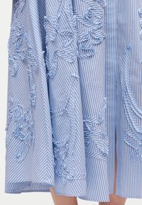 Φούστα με μπλε και λευκές ρίγες, με υφή υφάσματος που ανασηκώνεται και απαλά πιέτες, φορεμένη πάνω από γυμνά πόδια.