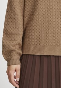 Pull en maille texturée marron avec un ourlet côtelé, présentant un motif en losange. Manches longues bouffantes se terminant par un poignet lisse et ajusté.