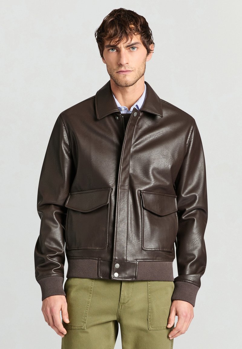 Mann trägt eine dunkelbraune Lederjacke mit aufgesetzten Taschen vorne, gerippten Bündchen und Bund, über einem hellen Hemd und olivgrünen Hosen.