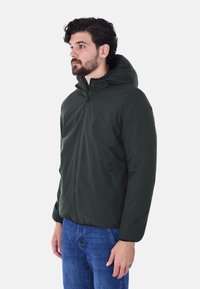 Giacca con cappuccio verde scuro, chiusura frontale con zip, polsini elastici e tasche laterali, realizzata in materiale liscio e resistente all'acqua.