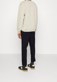 Man som bär en beige långärmad sweatshirt, svarta byxor, mörka strumpor och svarta sneakers med vit "PS"-logotyp på hälen, stående med ryggen vänd mot kameran.