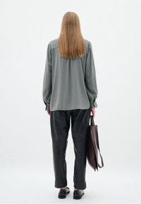 Blusa a maniche lunghe di un verde smorzato con polsini arricciati, abbinata a pantaloni di pelle nera. La persona tiene una borsa a tracolla marrone.