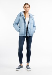 Lichtblauwe pufferjack met capuchon, ritssluiting aan de voorkant en zijzakken, gedragen over een witte top en donkere skinny jeans. Casual schoeisel maakt de look af.