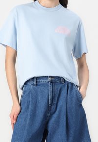 Vaaleansininen puuvillapaita, jossa on pyöreä kaula-aukko, lyhyet hihat ja vaaleanpunaista graafista tekstiä vasemmassa rinnassa, yhdistettynä korkeavyötäröisiin farkkuroihin.