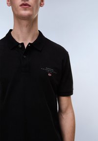 Jeune homme portant un polo noir Napapijri avec des boutons et un petit logo sur la poitrine, sur un fond clair uni.