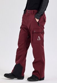 Burgundyfärgade utomhusbyxor gjorda av slitstarkt material, med sidofickor, svart accent vid fållen och en logotyp på övre låret.