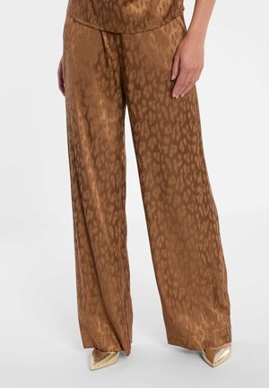 Braune, weit geschnittene Hosen mit dezentem Animalprint, kombiniert mit goldfarbenen Schuhen mit spitzer Spitze, Nahaufnahme des Unterkörpers und der Hand.