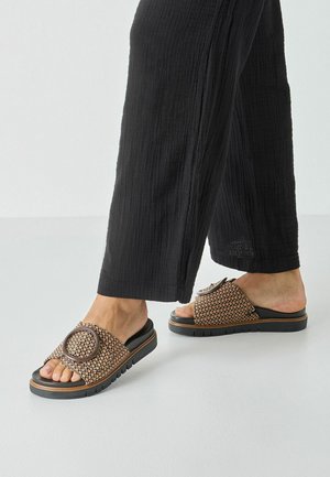 Woven brown slide sandals with a circular accent, black soles, and textured straps, paired with black wide-leg pants.