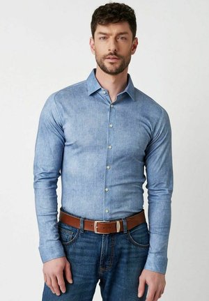 Mann mit kurzen lockigen Haaren, der ein tailliertes hellblaues Hemd mit Knöpfen, blaue Jeans und einen braunen Gürtel vor einem schlichten Hintergrund trägt.