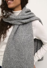 Écharpe en laine grise tricotée avec un motif en chevrons, douce au toucher. Elle est drapée autour du cou et présente une apparence texturée et chaleureuse.